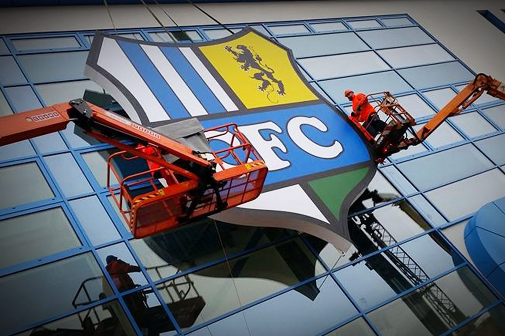 Großes Vereinslogo wird mit Hebebühnen an Glasfassade montiert