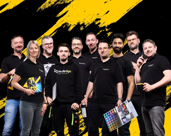 Das Team von sw/de/sign in schwarzer Arbeitskleidung vor einem dynamischen gelb-schwarzen Hintergrund – mit Farbmustern, Technik und guter Laune