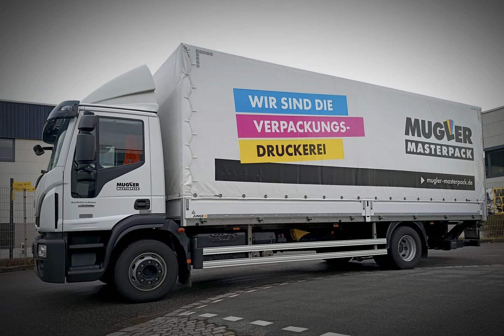Weißer LKW mit bedruckter Plane und großflächiger Werbebeschriftung für Mugler Masterpack auf einem Firmengelände.