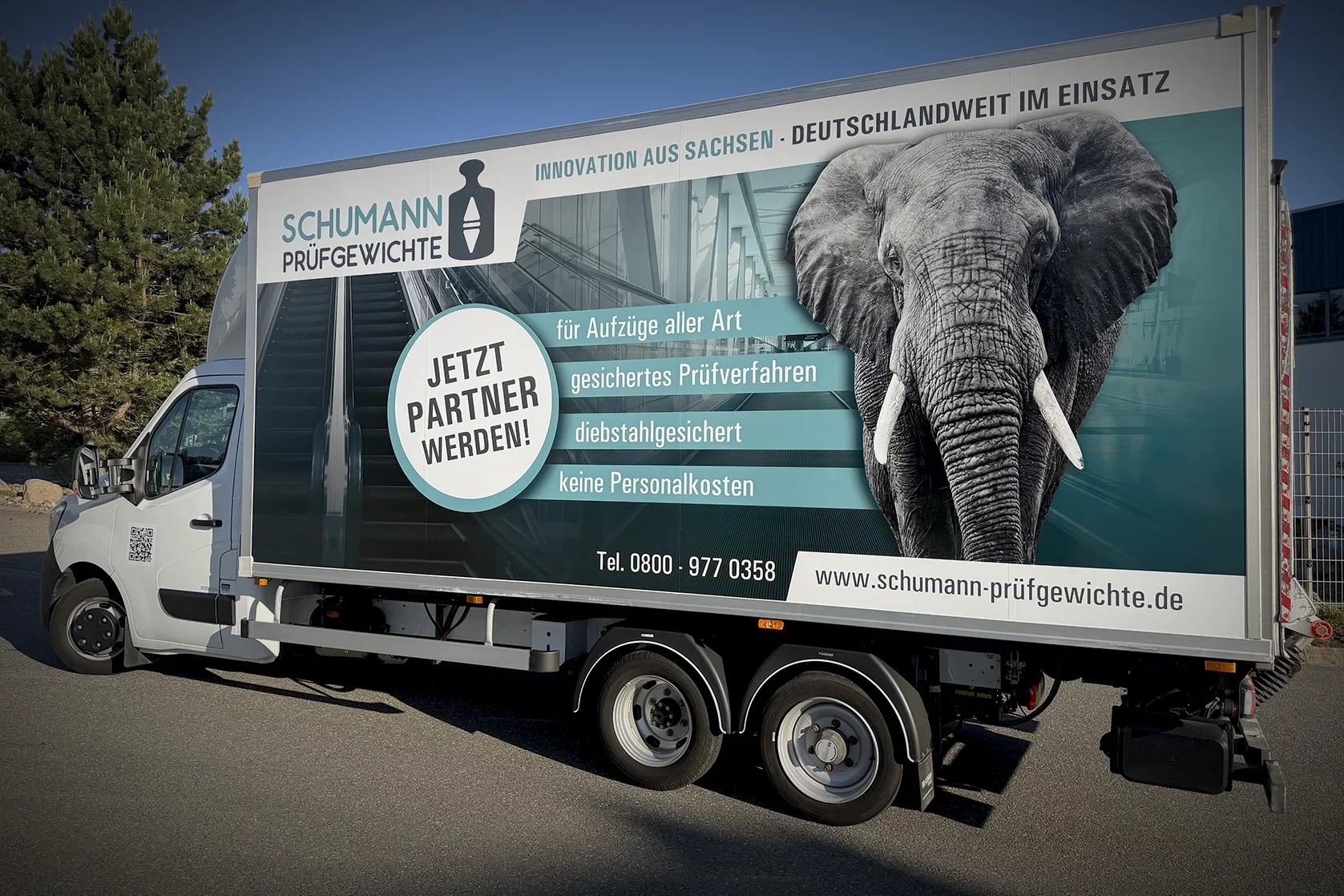 LKW-Planenbeschriftung für Schumann Prüfgewichte mit großformatigem Elefantenmotiv, Unternehmenslogo und Leistungsbotschaften zur Aufzugsprüfung auf einem Anhängerfahrzeug.